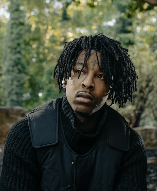 Headshot of the rapper 21 Savage slouching in the woods